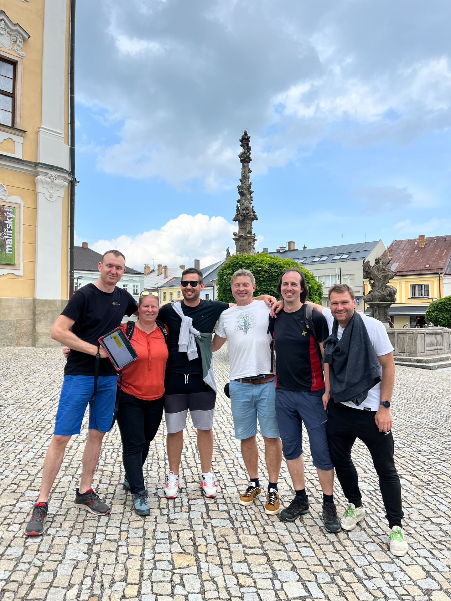 Six people pose with a tablet on a cobblestone square in front of a historic stone column