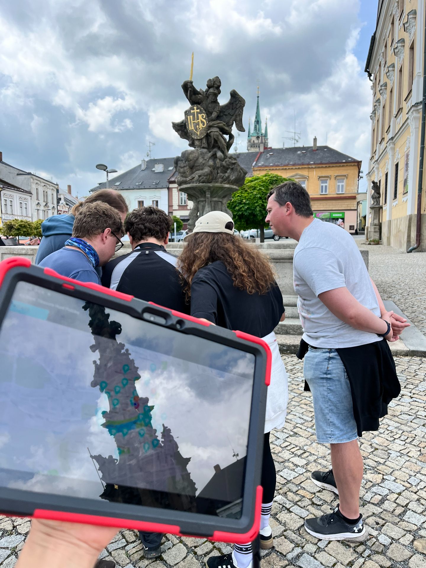 Person holds a tablet with a map in front of a group gathered by a fountain on a town square